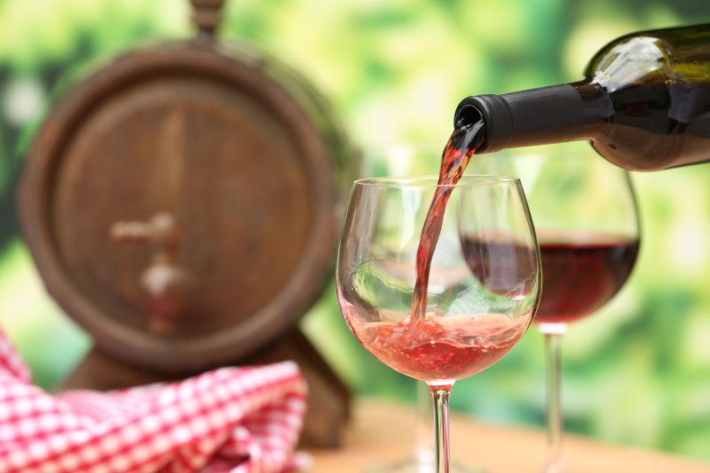 有木酒桶的绿色户外背景下向高脚杯中注入红酒