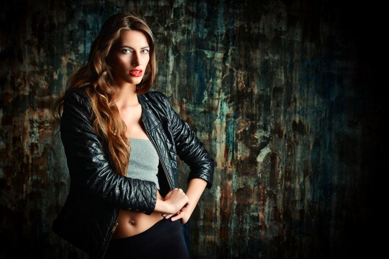 站在黑暗的老旧墙前的穿着黑色皮夹克的美女