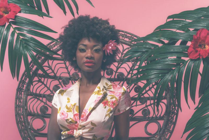 在粉红色背景上性感的女高手坐在棕榈叶之间的柳条椅上