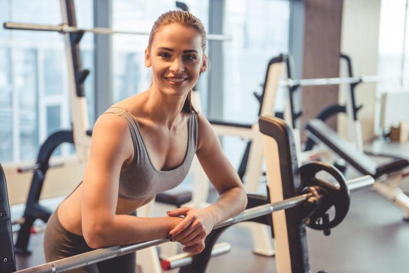 靠在杠铃上看着相机微笑着的有魅力的年轻女子