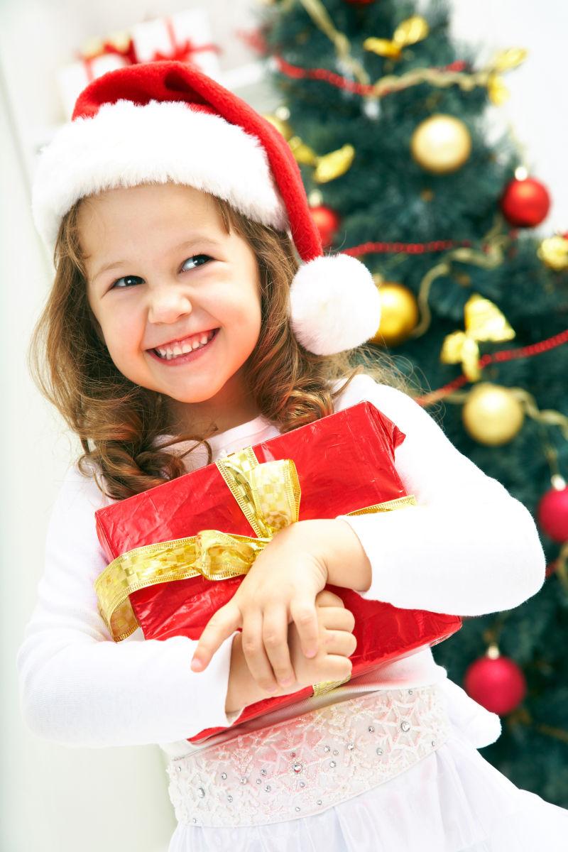 圣诞树前带着圣诞帽抱着礼物微笑的小女孩