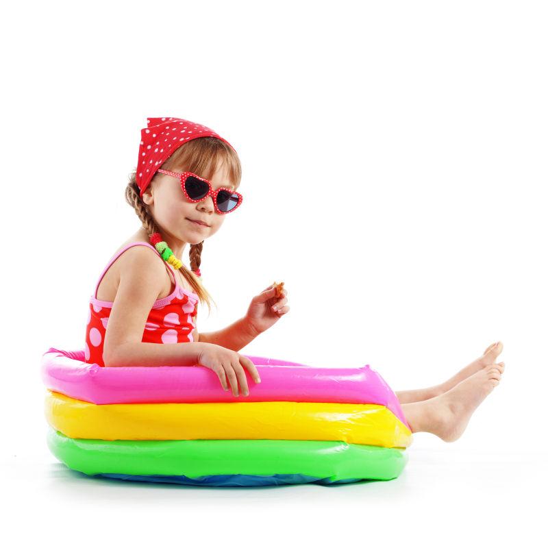 白色背景中带着太阳镜穿着泳衣坐在游泳圈中的小女孩