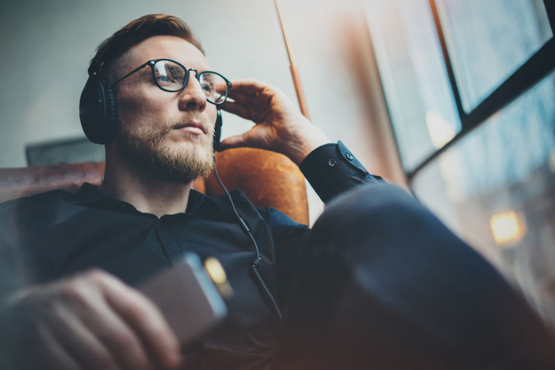 肖像:英俊的胡须男人戴着眼镜头戴耳机在现代化的家里听音乐男人坐在老式的椅子上手持智能手机放松全景窗户背景模糊