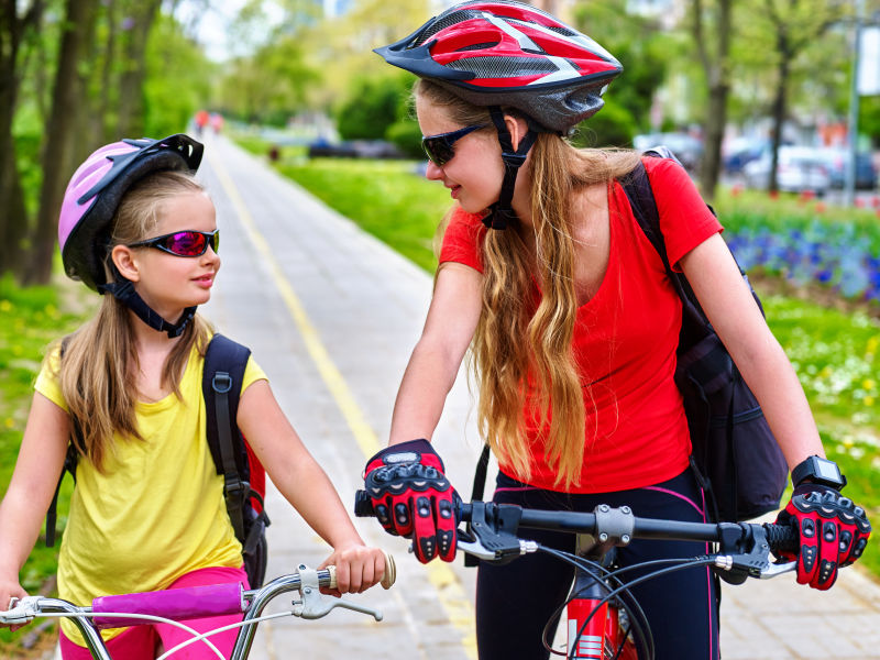 女孩们戴着自行车头盔和玻璃背着帆布包骑着自行车女童在黄色自行车道
