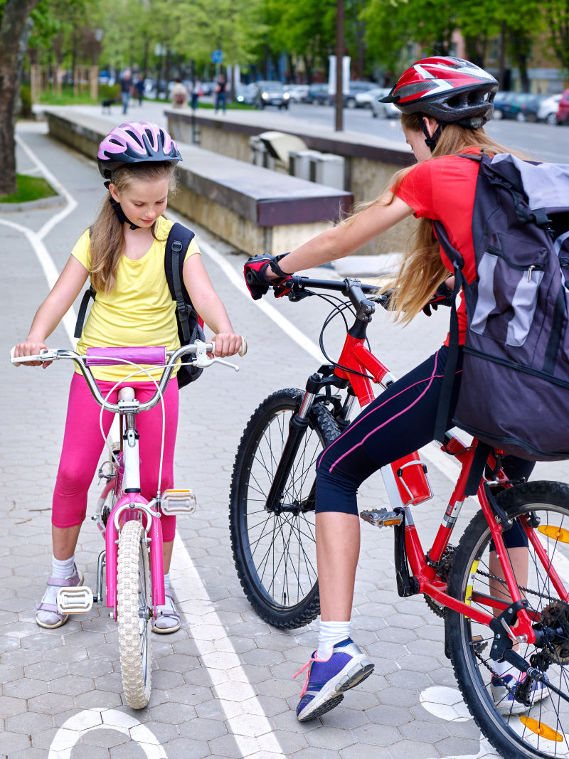 骑自行车的女孩女孩们戴着自行车头盔和玻璃背着帆布包骑着自行车女童骑自行车在白色自行车道上相遇自行车共享计划节省了城市街道的金钱和时间那里