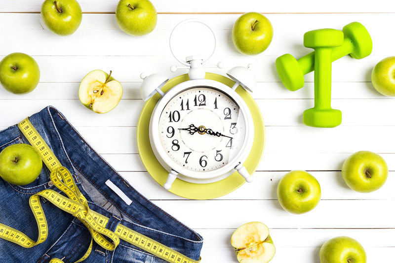 带卷尺的闹钟-蓝色女式牛仔裤-绿色苹果和白木背景的哑铃-健康的生活方式-减肥和减肥的概念