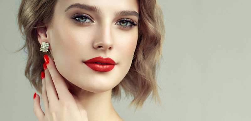 漂亮的模特儿卷发短嘴唇红指甲上的红色修指甲美容和美容护理金色头发的现代造型耳环珠宝和配件