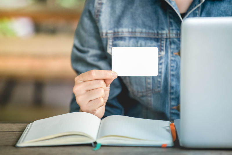 女性的Jean Jecket手拿白色名片-负责联络工作-空白纸卡模型-办公桌上有笔记本电脑和空白笔记本电脑的工作场所