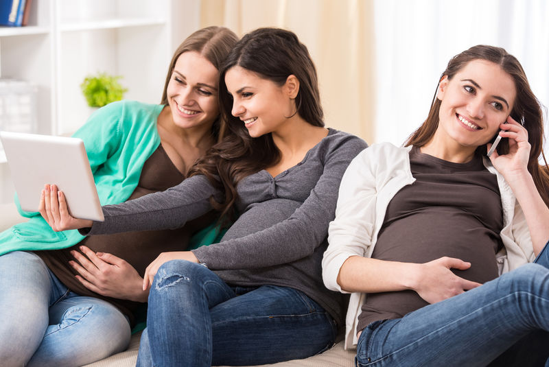 微笑的孕妇在网上看一些有趣的东西而她们的朋友正在打电话