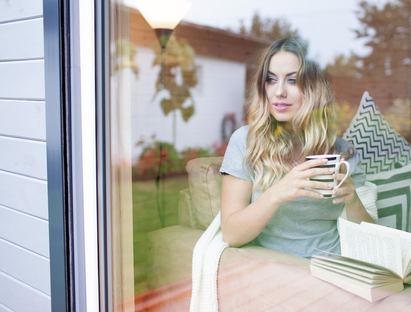 年轻漂亮的金发女郎端着一杯咖啡坐在窗边后院玻璃上的反光懒散的休假概念