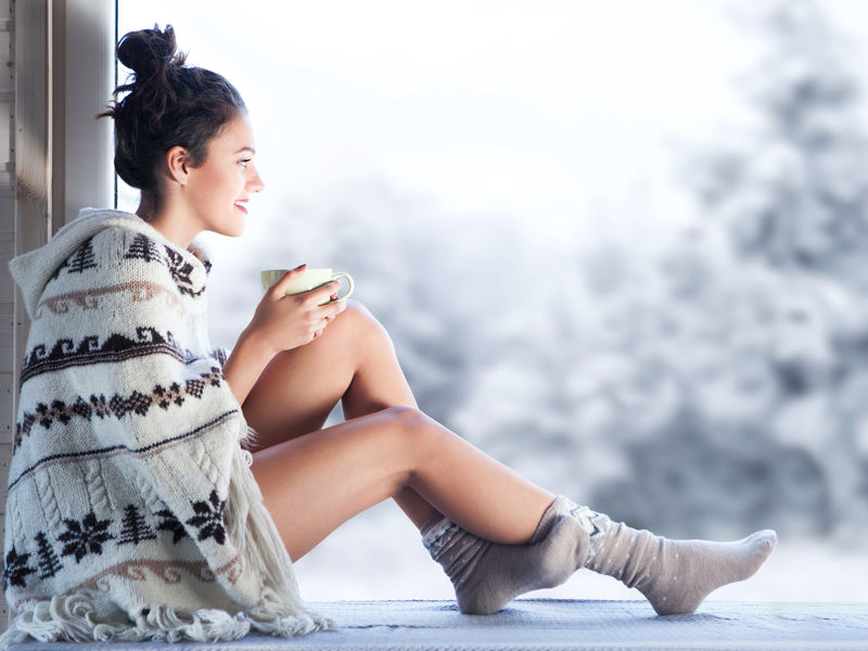 年轻漂亮的黑发女人喝杯咖啡坐在家里穿针织北欧打印雨披的窗口模糊的冬季雪树背景
