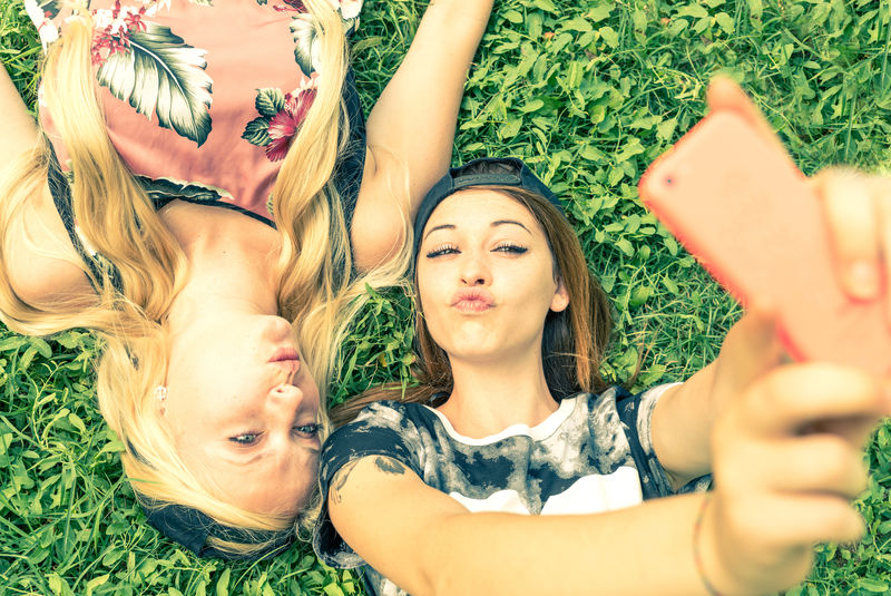 两个漂亮的女孩在自拍——穿着运动休闲装的年轻女子躺在草地上自拍时玩得很开心