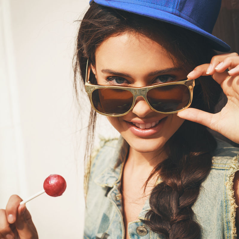 戴着蓝帽子和牛仔裤夹克的年轻性感女人透过太阳镜看镜头-户外-生活方式