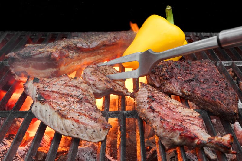 在烧烤架和叉子上混合烤好的肉-猪胸肉、排骨、牛排-背景中有火焰