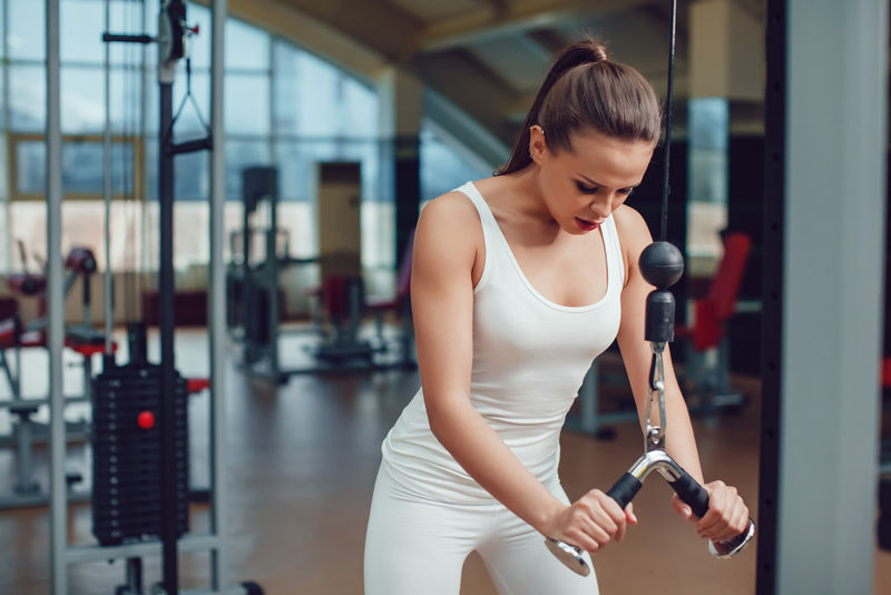 漂亮的女孩穿着白色的运动背心和紧身衣在健身房的模拟器上做运动。