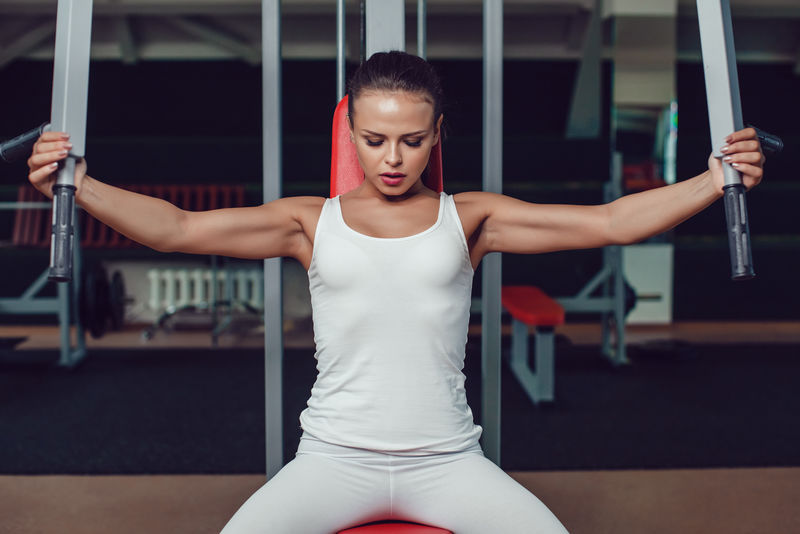 漂亮的女孩穿着白色的运动背心和紧身衣在健身房的模拟器上做运动。