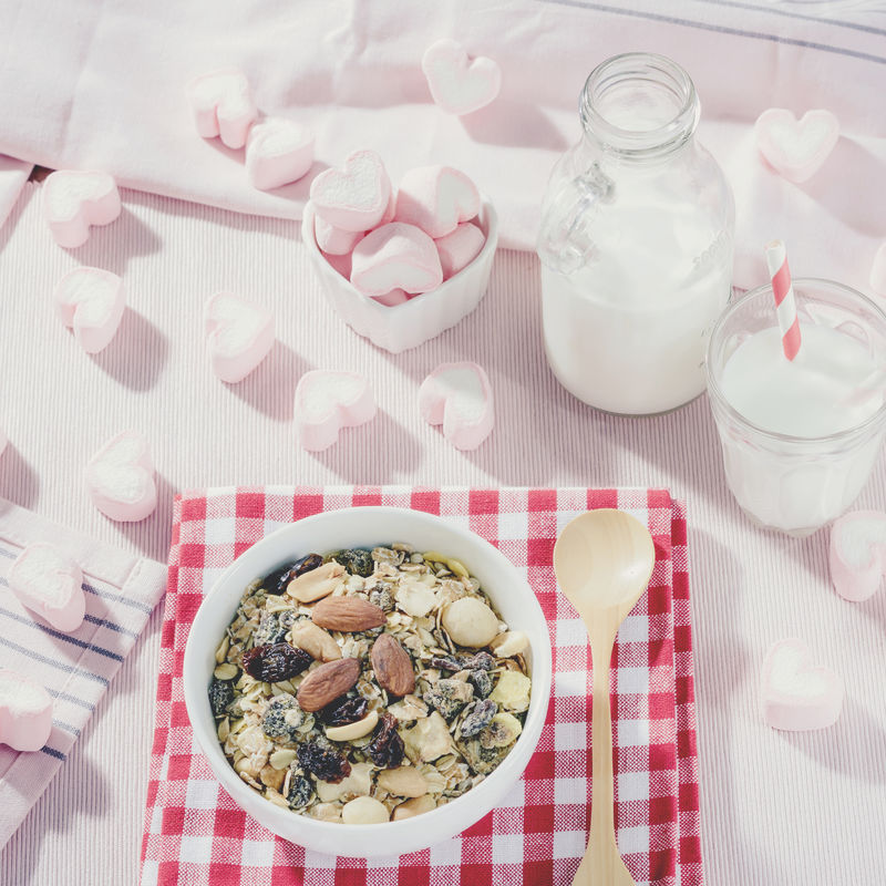 早餐套餐。将燕麦格兰诺拉麦片与粉红心形甜棉花糖混合在白色碗中，与白牛奶一起装在玻璃杯和瓶子里，俯视图，健康的食物理念，柔和的色调。