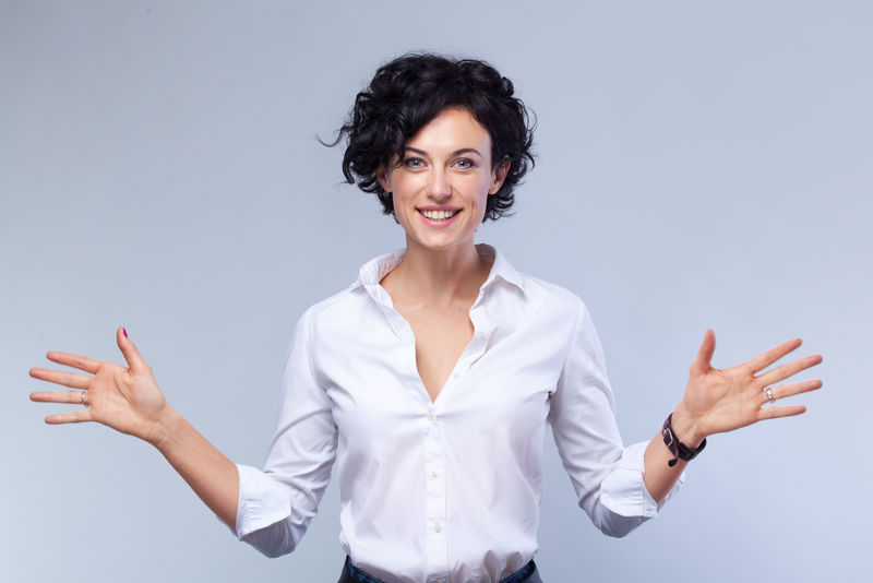 快乐的年轻黑发女,灰色背景卷发,复印空间,张开双臂看着相机。