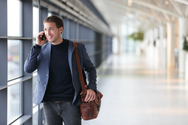 在机场里行走的城市商人用智能手机交谈-穿着西装夹克和肩包的休闲年轻商人-20多岁的英俊男模特