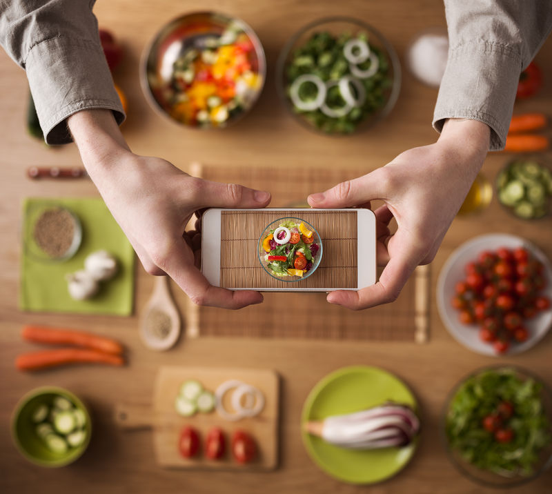 拿着智能手机的男人手特写镜头-厨房桌上的工作台背景是新鲜蔬菜和餐具