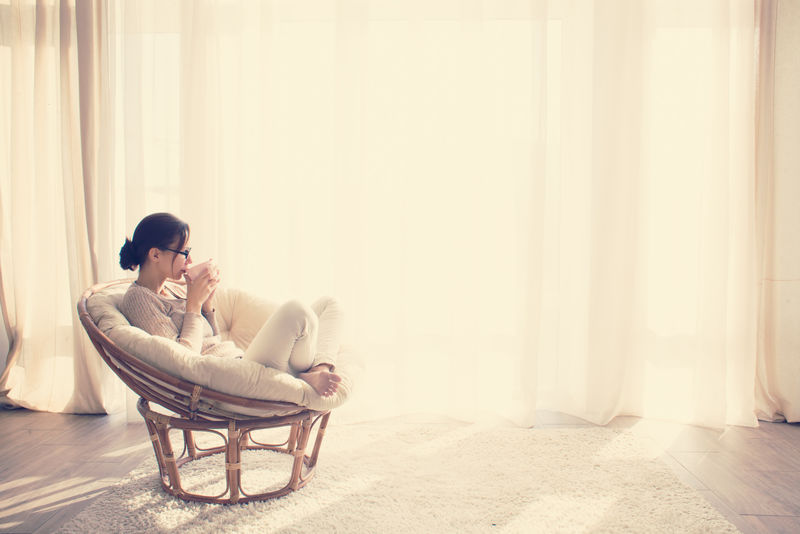 坐在窗户前的现代椅子上的年轻女性在起居室里放松地看书-喝咖啡或茶-喝Instagram Toning
