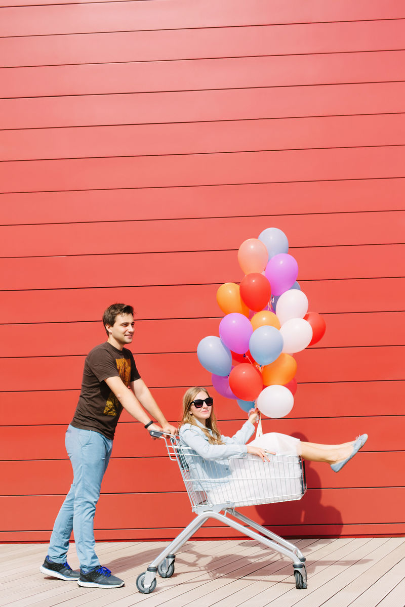 两个快乐的年轻人约会和娱乐的照片-女士坐在购物车里-手里拿着彩色气球-红色背景-户外