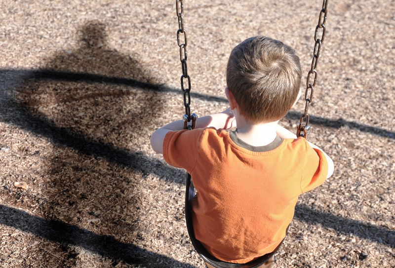 一个小男孩坐在秋千上-看着操场上一个男人或恶霸的影子-用于绑架、防御或安全概念