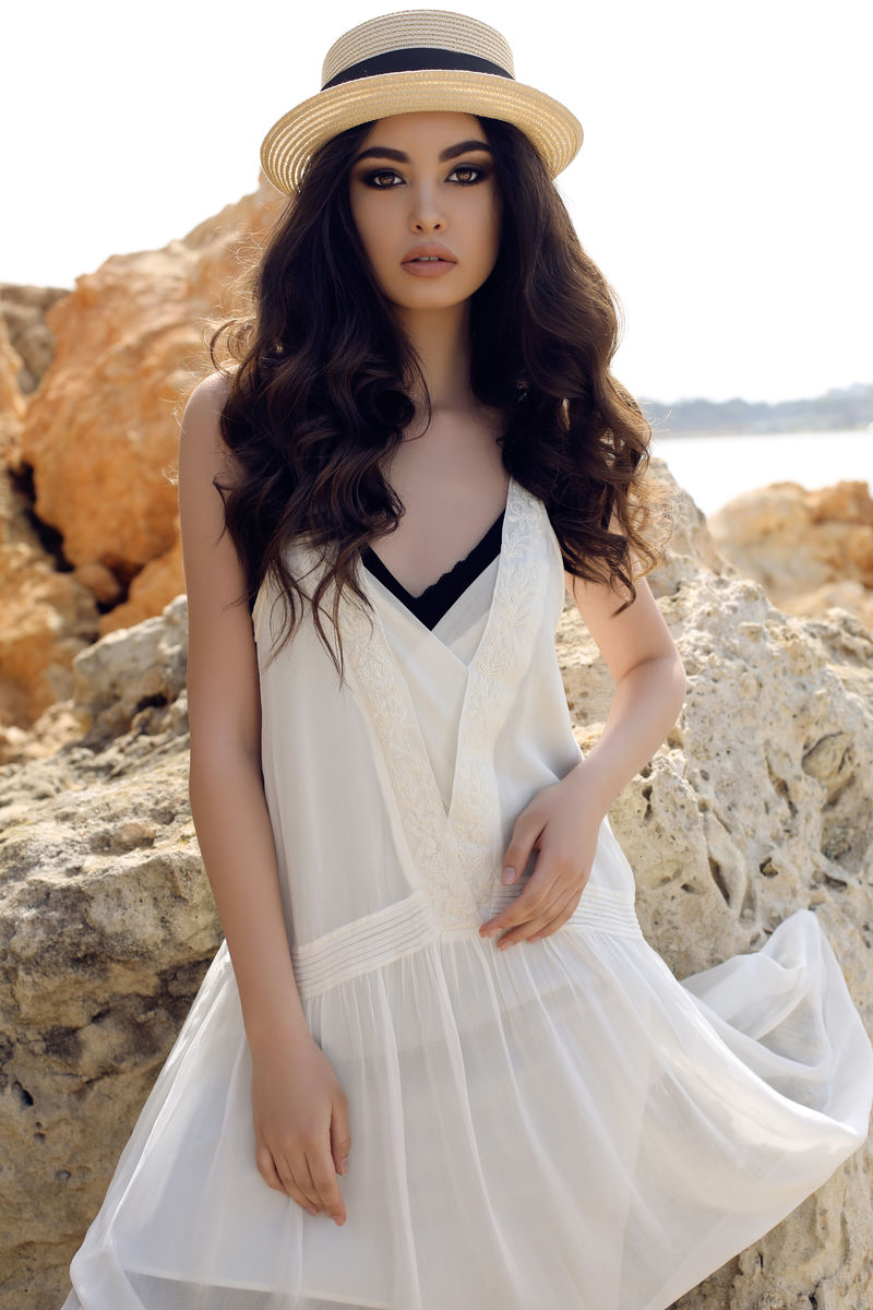 时尚户外照黑发美女身穿休闲优雅服装戴帽子在夏日海岸摆造型