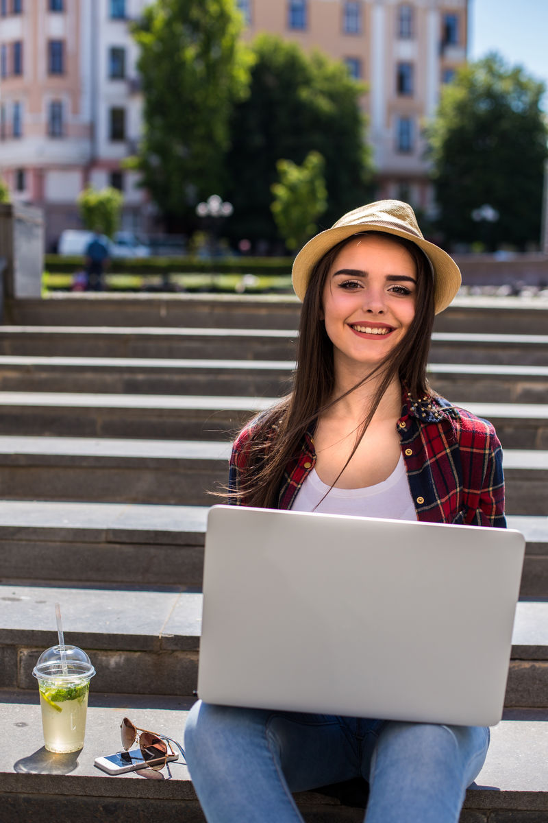 一位快乐的年轻女子坐在城市的楼梯上，在户外用笔记本电脑看镜头