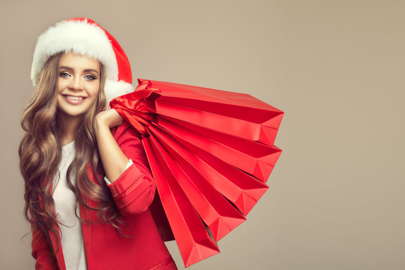 圣诞帽上可爱微笑的女人的肖像-手里拿着红色的购物袋-圣诞节