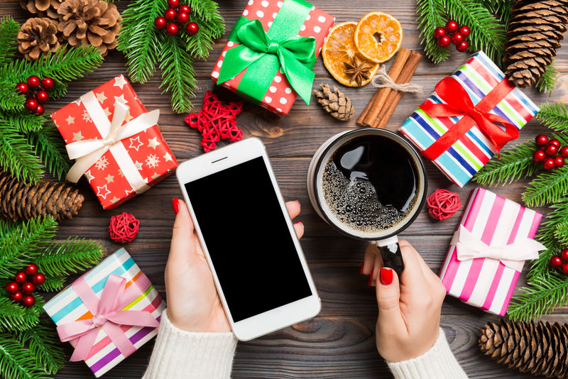一个女人一手拿着一杯咖啡，另一手拿着一部手机的俯视图。冷杉和圣诞装饰品。新年假期概念。模型