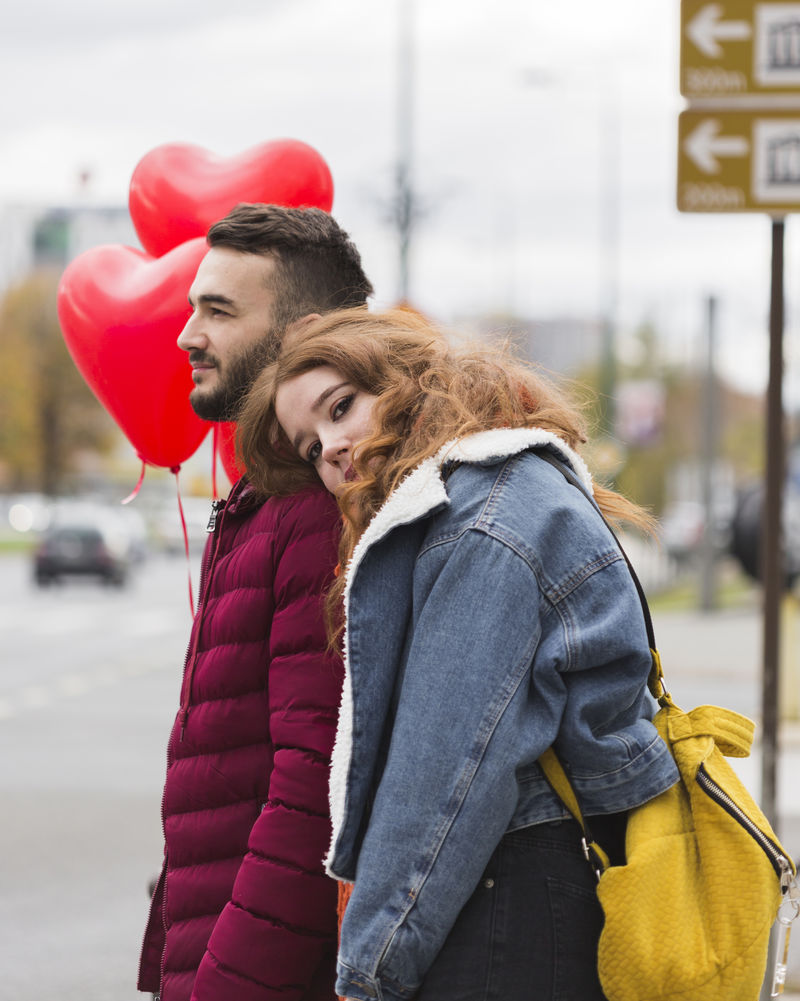 一对笑容可掬的异族夫妇走在室外的停车场上-白人握着他的黑人女友的手-她手里拿着一顶深红色的帽子-他们穿着休闲风格的衣服-水平方向