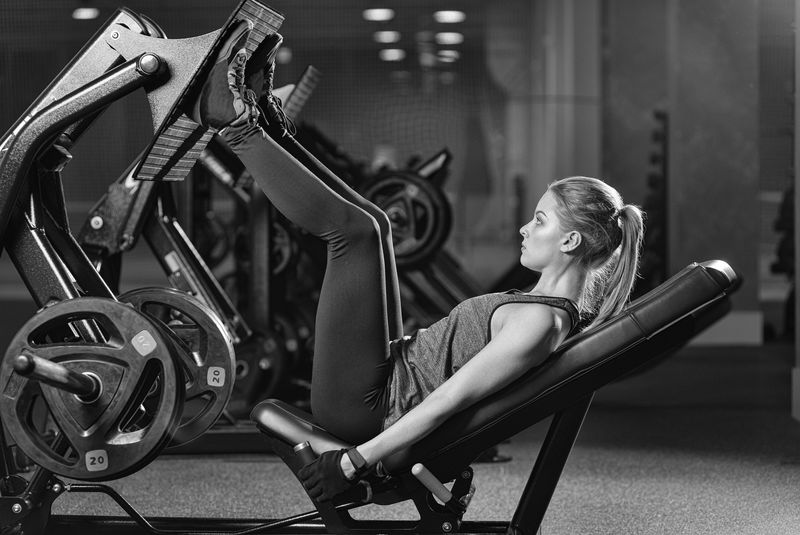 运动健美女子在健身房使用腿部压重机。漂亮的黑发女人在模拟器里锻炼。在机器上训练她的四头肌。黑白相间。