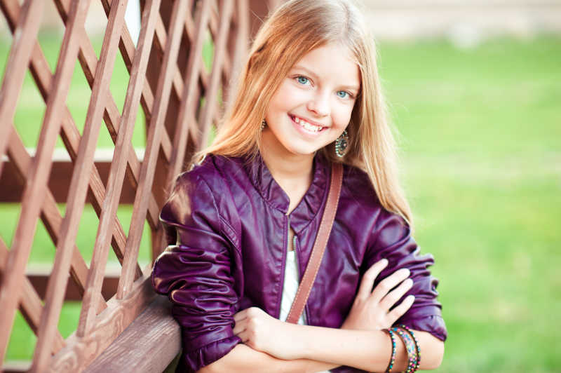 在绿色的自然背景下的穿着时髦外套的微笑的金发女郎看着相机