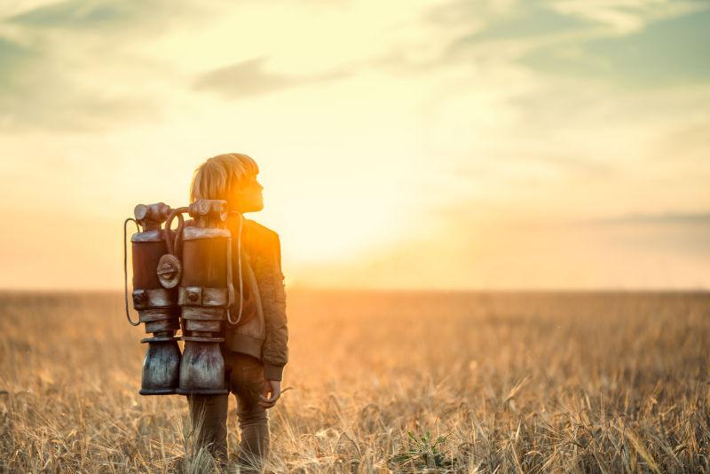 日落下背着喷气式飞行器站在田野里的男孩