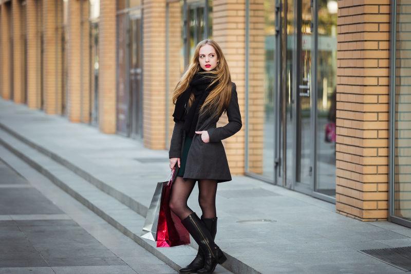 黄色建筑前穿着黑色风衣拎着购物袋的美女